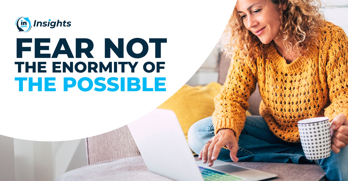 A promotional graphic from 'in/sync Insights' with the text 'FEAR NOT THE ENORMITY OF THE POSSIBLE.' prominently displayed in bold, blue font. The image features a woman sitting on a couch, engrossed in work on her laptop. Her curly blonde hair partially obscures her face as she appears to be deeply concentrated. The setting includes a brightly colored cushion with floral patterns, suggesting a casual and creative work environment. The graphic design is modern and motivational in style.